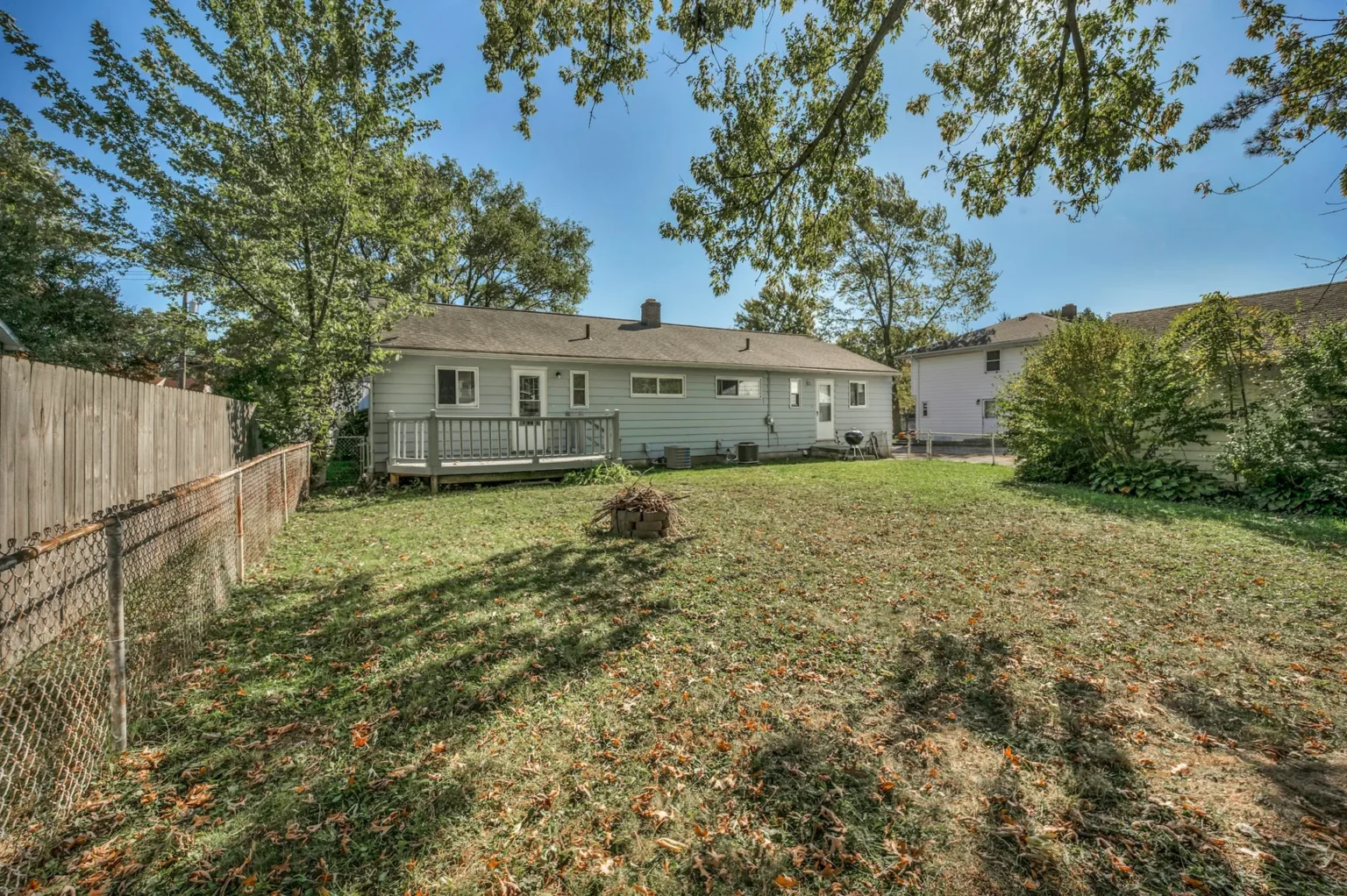A backyard lawn with fallen leaves under mature trees whose branches reach over a single story house
