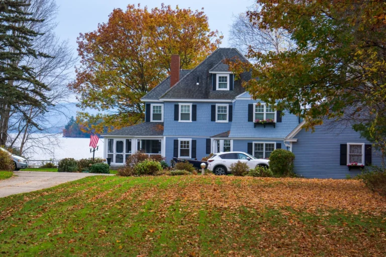Tall maples and oaks in orange and gold fall color behind a blue shingled house and a leaf covered lawn, a setting for tree removal in Edgewater