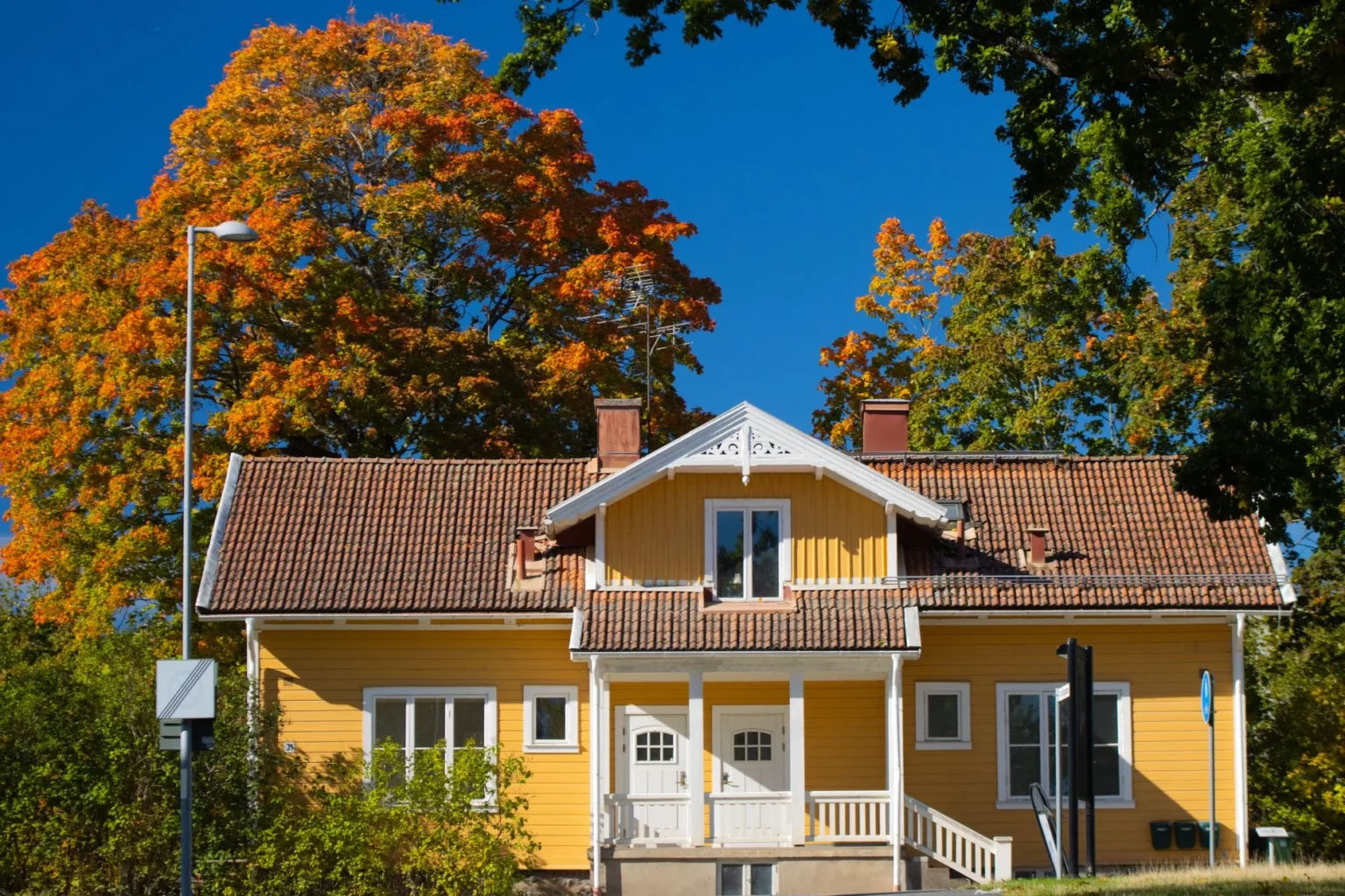 Large maple with orange autumn leaves behind a yellow two story house with a tiled roof